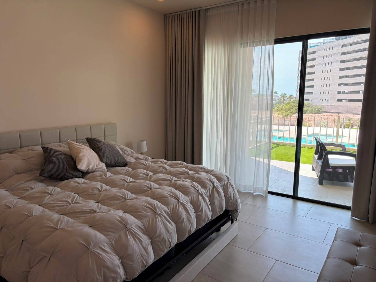 A bedroom with a large bed, a nightstand, a lamp, and a view of a swimming pool through a glass door.