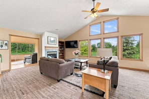 A spacious living room with a fireplace, a ceiling fan, and large windows offering a view of the outdoors. - Acres Away - 50 Acre Farmland, Family Retreat, Private Yard (Fennville)