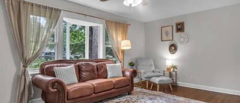 Una sala de estar con un sofá de cuero marrón, un tapete estampado, un ventilador de techo y una ventana con cortinas.