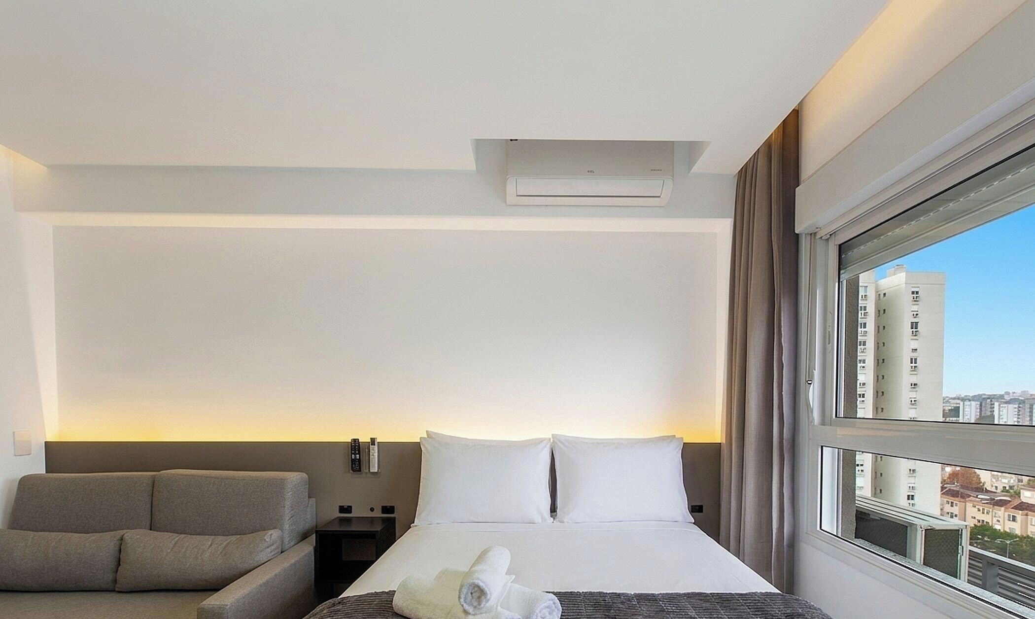 Quarto de hotel com cama, sofá, janela com vista para a cidade e ar condicionado fixado na parede.