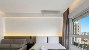 Quarto de hotel com cama, sofá, janela com vista para a cidade e ar condicionado fixado na parede.