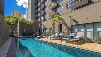 Uma piscina com espreguiçadeiras e guarda-sóis, cercada por um edifício com varandas e janelas.