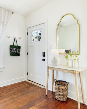 Un couloir avec une porte blanche, un miroir avec un cadre doré, une petite table avec une lampe et un panier en osier.