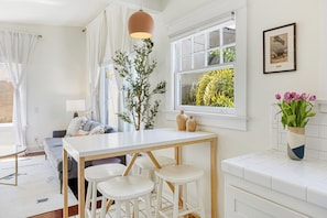 A modern kitchen with a white countertop, wooden stools, a hanging pendant light, and a window with a view of greenery. - Cozy 1-bedroom apartment in beautiful Venice Beach with AC (Los Angeles)