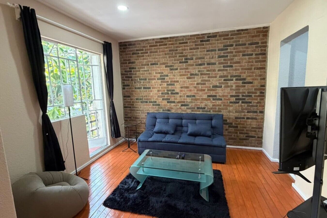Un salon avec un mur en briques, un canapé bleu, une table basse en verre, un tapis noir et un téléviseur.