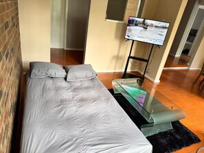 A single bed with a grey duvet, a glass table with a black furry rug, and a television mounted on the wall displaying a snowy scene. - Amplio Oasis con Balcón en Roma Norte (Mexico City)