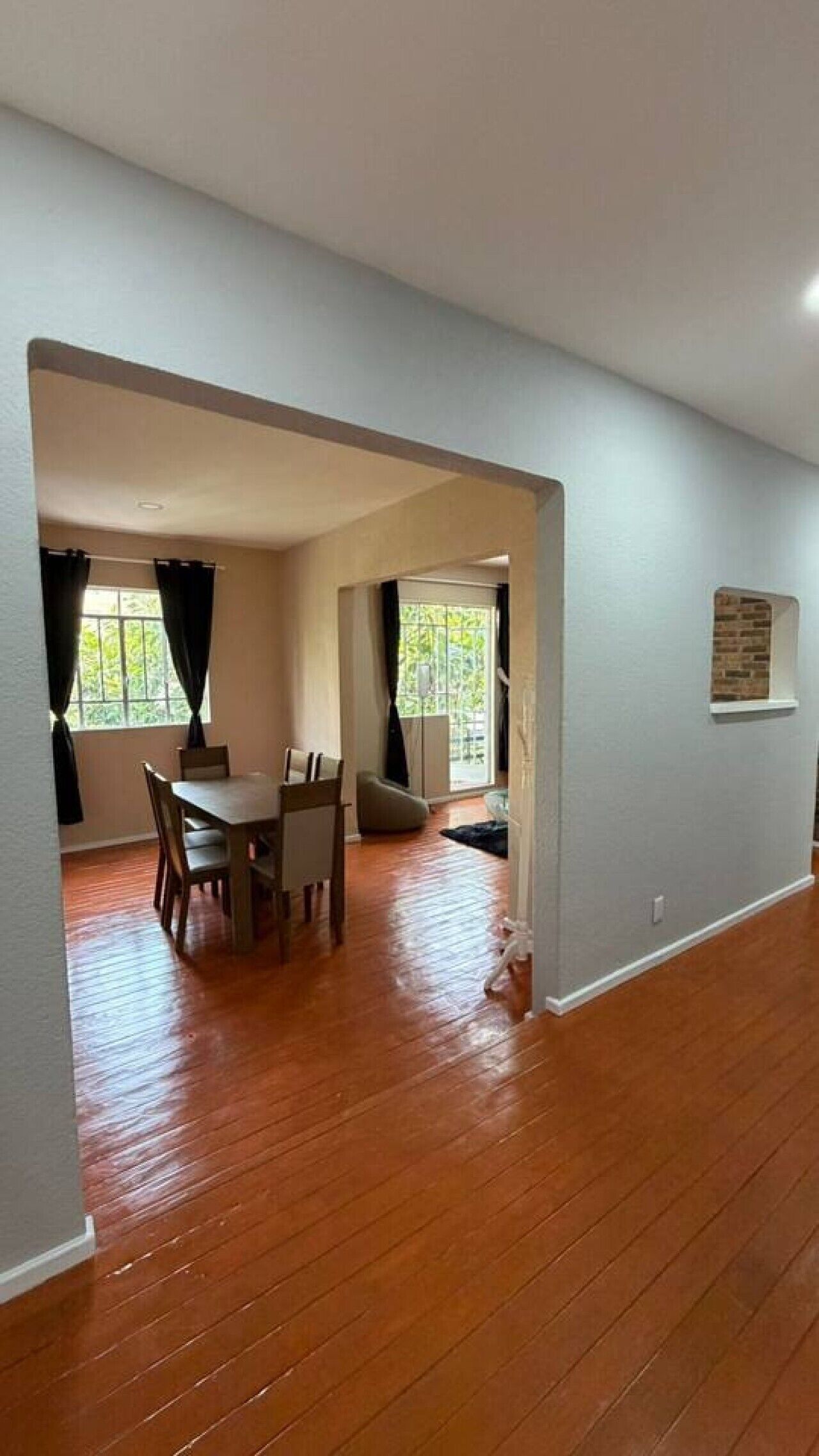 A spacious room with a wooden floor, a dining area with a table and chairs, and a window with curtains.
