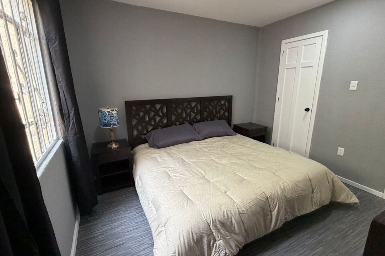 A bedroom with a bed, nightstands, a lamp, a window with curtains, and a door.