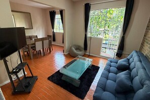 A living room with a sofa, a glass coffee table, a TV, a dining area with chairs, and a view of greenery through a window. - Amplio Oasis con Balcón en Roma Norte (Mexico City)