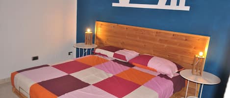A bedroom with a wooden headboard, a bed with a multicolored quilt, two bedside tables with lamps, and a blue accent wall with a white shelf.