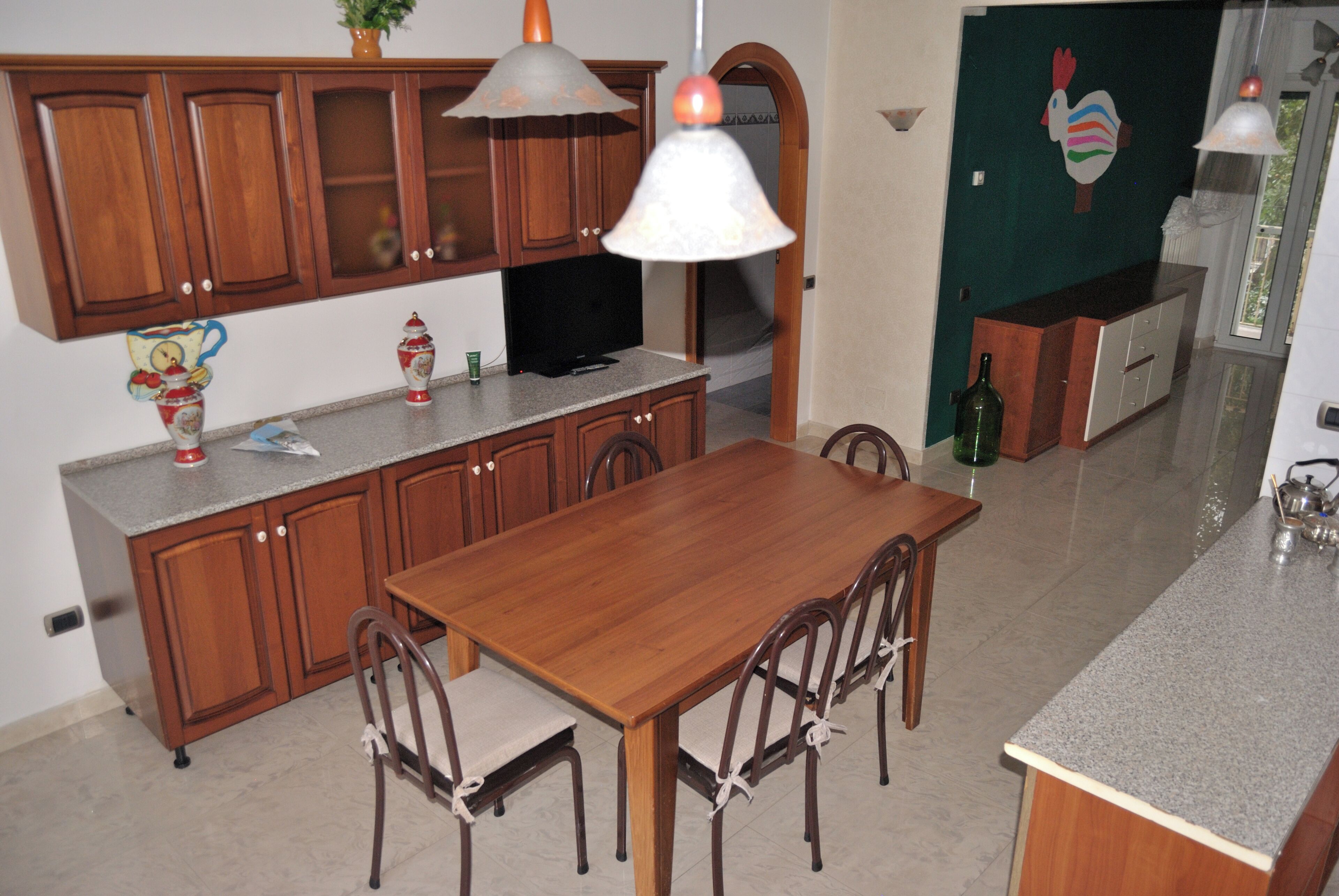 Une cuisine avec des meubles de rangement en bois, une table à manger avec des chaises, un téléviseur et un élément décoratif mural.
