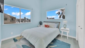 Quarto com cama, mesa de cabeceira, luminária e janela com vista para prédios e palmeiras.