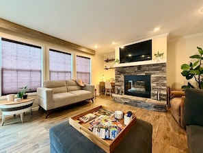 A living room with a fireplace, a sofa, a coffee table with magazines, and a dining table with a plant. - Mountain Top Farmhouse Retreat /Hot Tub/Arcade (Cosby)