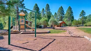 A playground with a slide and climbing structures, surrounded by trees and a gravel path.