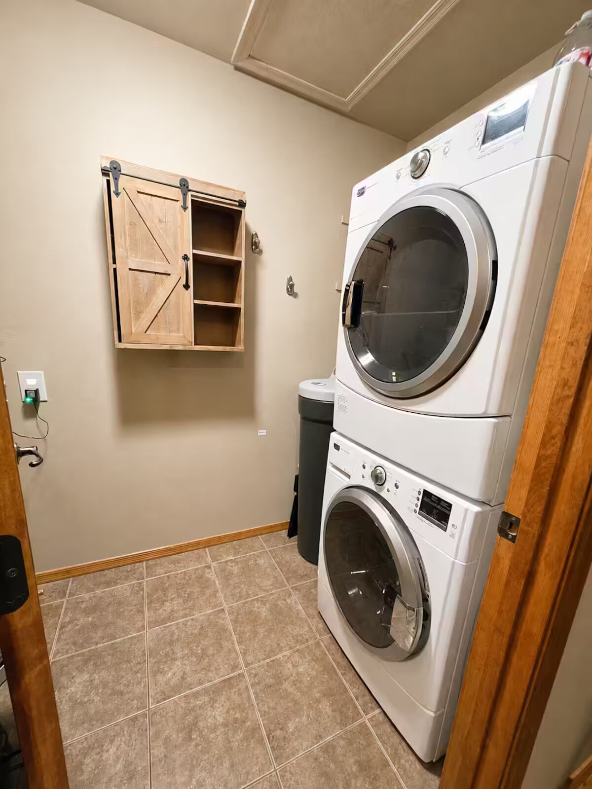 A laundry room with a stacked washer and dryer, a wooden cabinet with shelves, and a trash can.