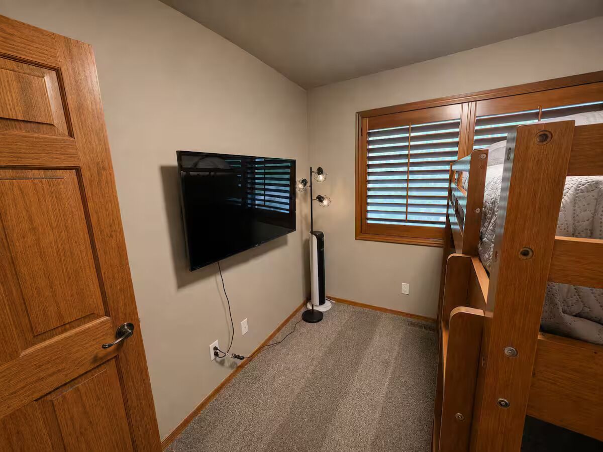 A room with a wooden door, a mounted television, a standing fan, and a window with blinds.