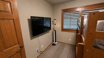 A room with a wooden door, a mounted television, a standing fan, and a window with blinds.