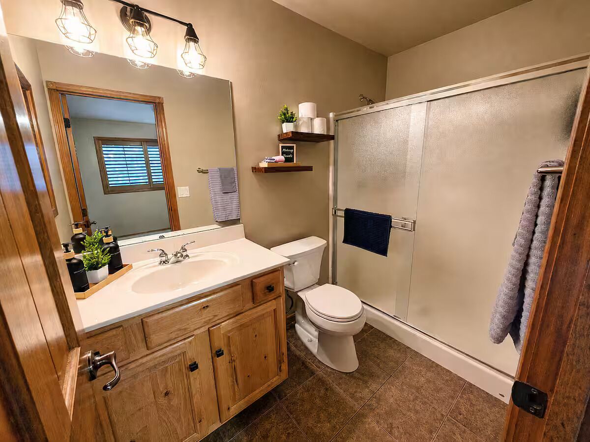Un baño con tocador de madera, inodoro blanco, cabina de ducha de vidrio y espejo con luces.