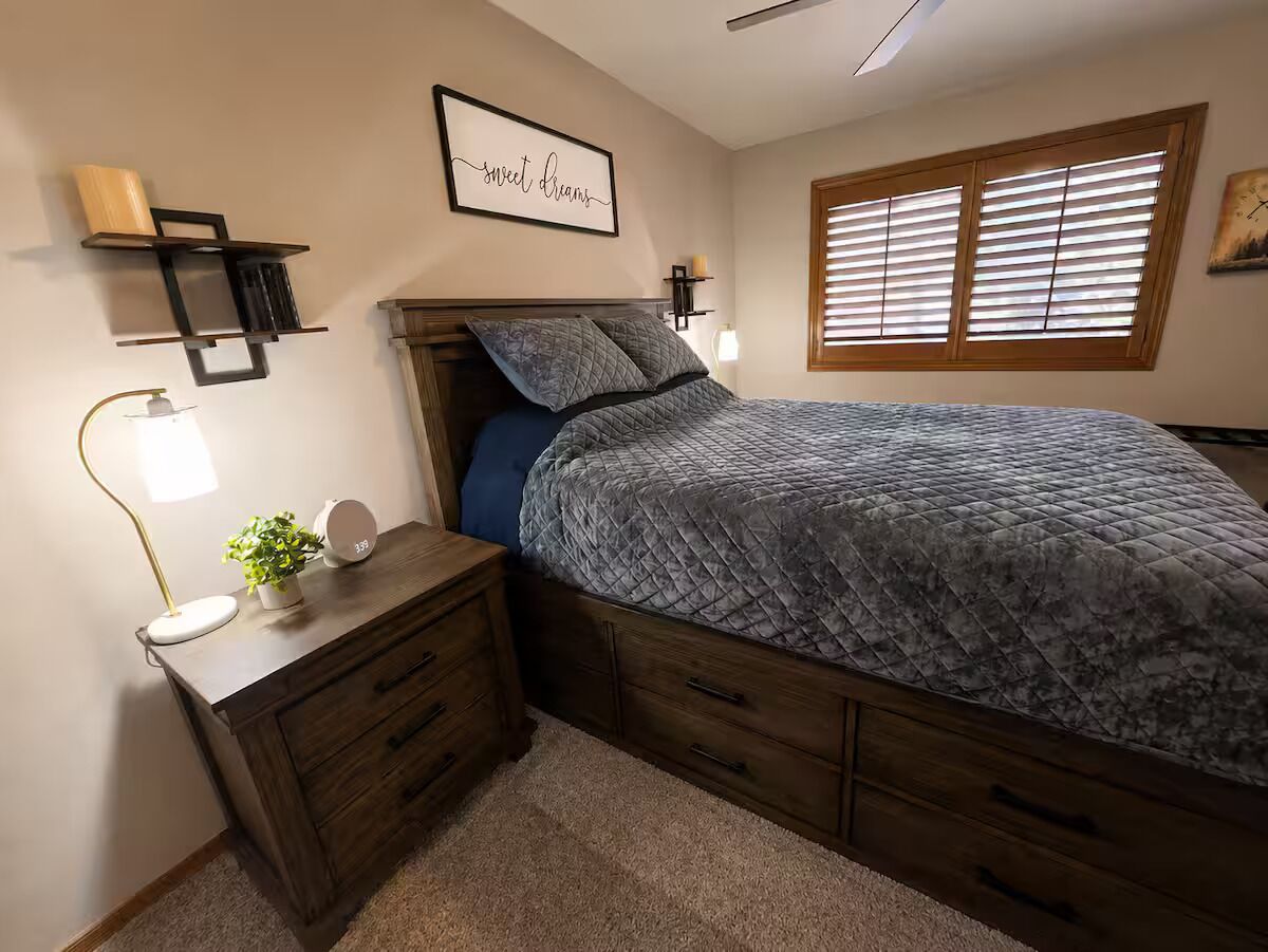 Une chambre à coucher comprenant un lit, des tables de chevet, une étagère fixée au mur, une lampe de chevet et une fenêtre avec des stores.