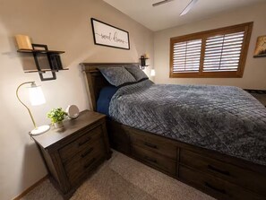 Une chambre à coucher comprenant un lit, des tables de chevet, une étagère fixée au mur, une lampe de chevet et une fenêtre avec des stores.