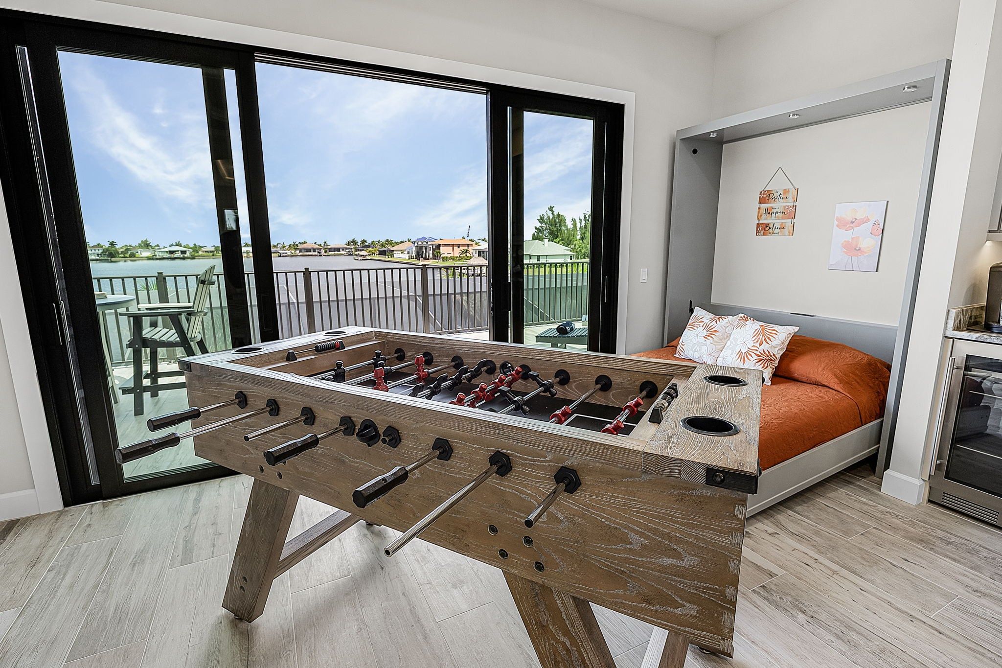A room with a foosball table, a bed with orange bedding, and a balcony with a view of the water.