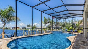 A swimming pool with a curved edge, surrounded by a covered patio area with chairs and a view of the water. - Waterfront Fun, Private Beach, Pool, Spa & Games - Laguna Lake House - Roelens (Cape Coral)