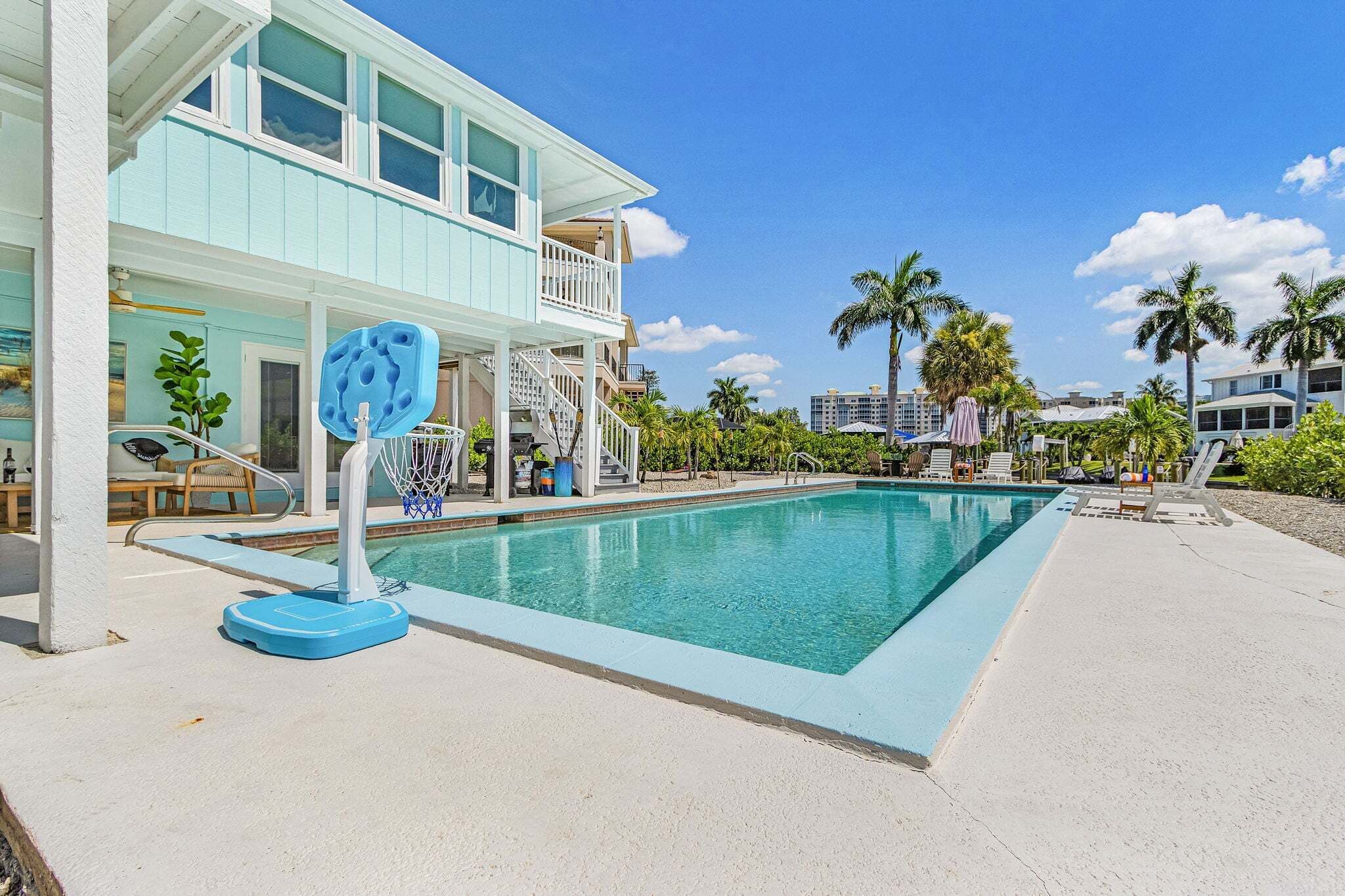 A swimming pool with a basketball hoop, a two-story building with a balcony, and palm trees in the background.
