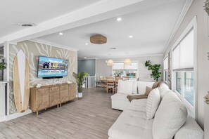 A spacious living room with a television mounted on the wall, a dining area with a table and chairs, and a large window providing natural light. - Gulfside Fun, Pool Days & Waterfront Adventures Await - Coastal Oasis - Roelens (Fort Myers Beach)