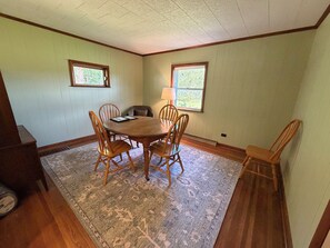 A dining room with a round wooden table, six wooden chairs, a rug, two windows, and a lamp.