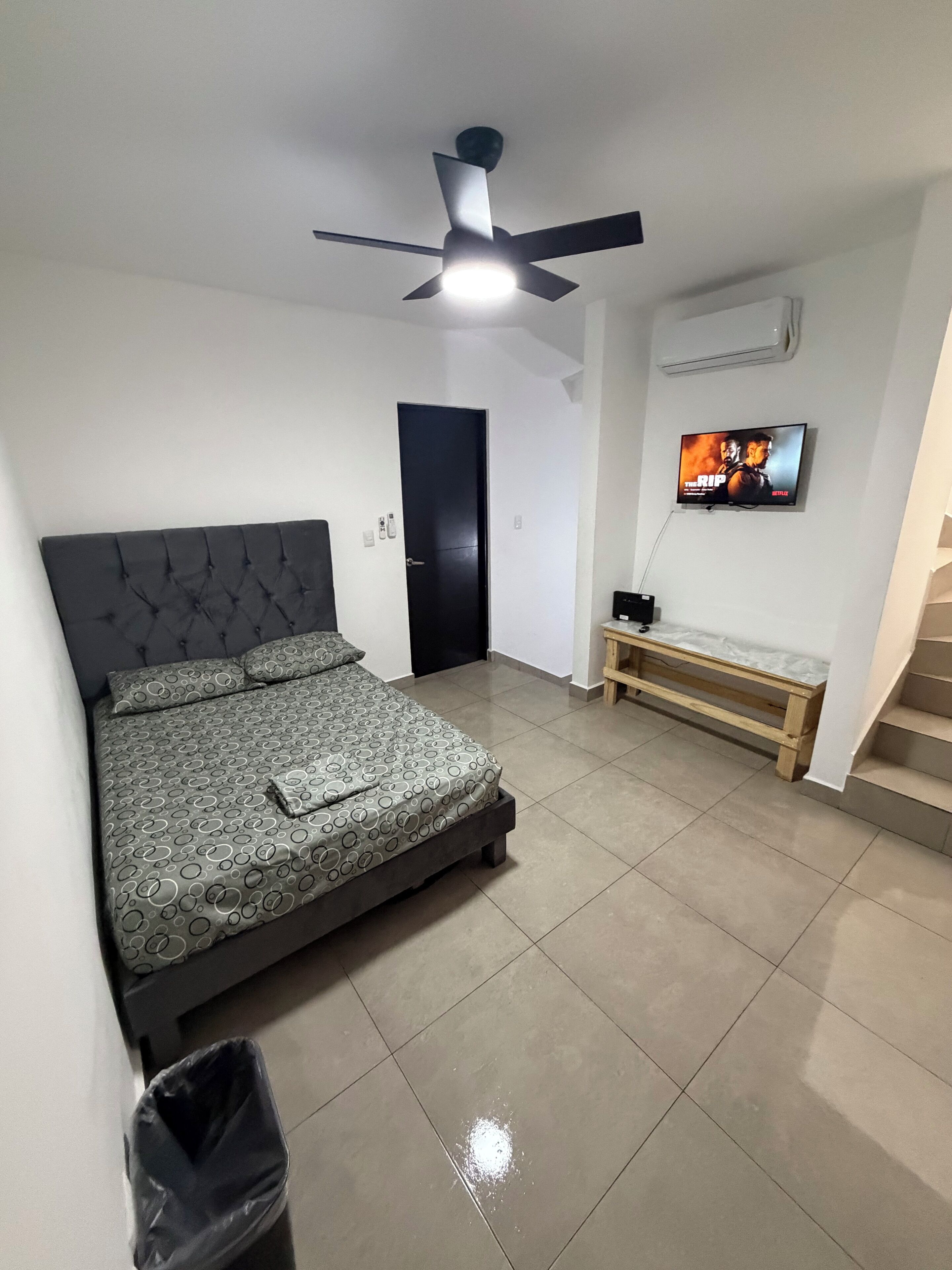 Een slaapkamer met een bed, plafondventilator, televisie, bankje en een deur.