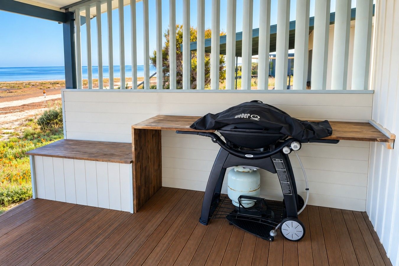 A portable grill with a gas tank and a bag on a wooden deck with a view of the beach and ocean.
