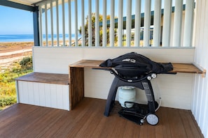 En grill til at tage med, med gasbeholder og taske, placeret på en træterrasse med udsigt til strand og hav.