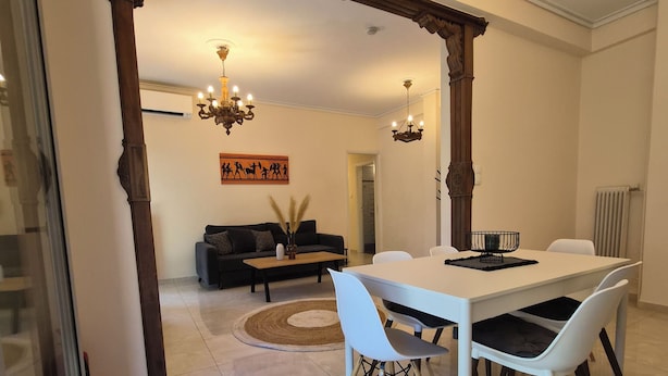 Ein Essbereich mit einem weißen Tisch, weißen Stühlen und einer Chandelüster. Im Hintergrund befindet sich eine Couch und ein Couchtisch.