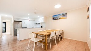 A modern kitchen with white cabinets, a central island, and a dining area with a wooden table and white chairs. - Breakers 5/5- Stone Real Estate Yamba (Yamba)