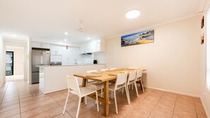 A modern kitchen with white cabinets, a central island, and a dining area with a wooden table and white chairs. - Breakers 5/5- Stone Real Estate Yamba (Yamba)