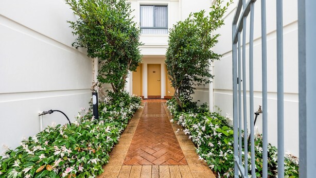 A corridor with a herringbone patterned floor, flanked by lush greenery and white flowers, leading to a door with a window above. - Breakers 5/5- Stone Real Estate Yamba (Yamba)