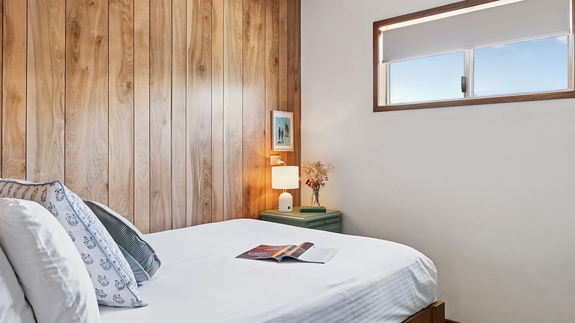 Een net opgemaakt bed met een witte laken en kussens, een nachtkastje met een lamp en een vaas gedroogde bloemen, en een raam met uitzicht op de lucht.