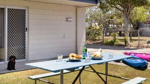 An outdoor patio with a picnic table set up, featuring a bottle, bowls of fruit, and glasses. - Brooms Head Bungalow (Brooms Head)