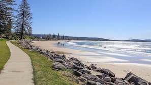 A coastal walkway with a sandy beach, a clear sky, and a line of trees.