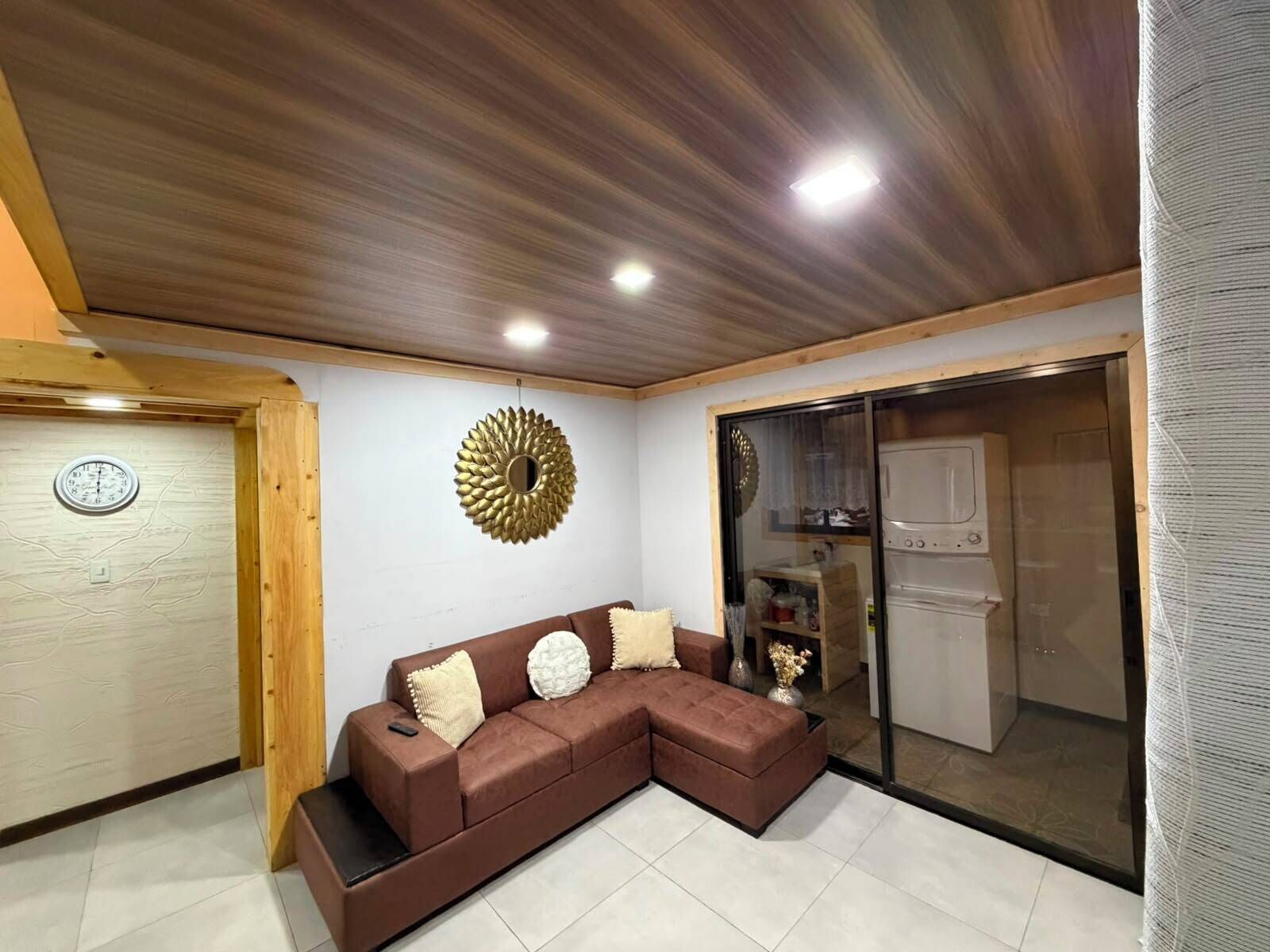 A modern living room with a brown sectional sofa, a decorative wall piece, and a kitchen area visible through a glass door.