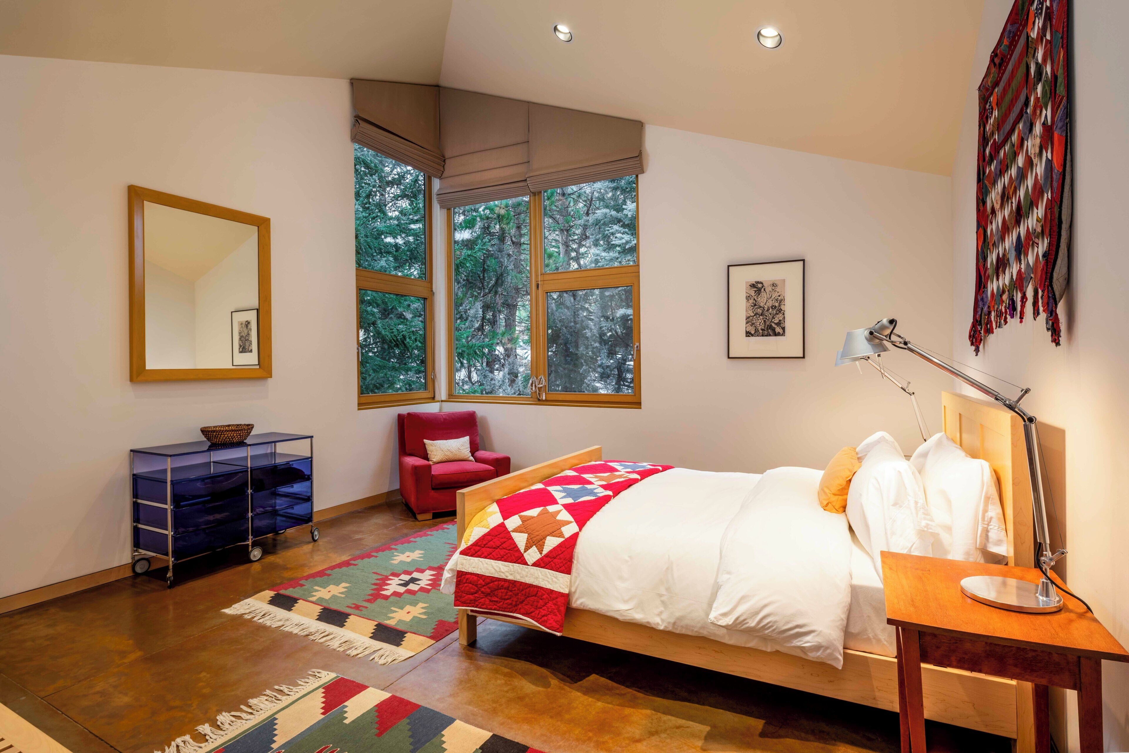 A bedroom with a large bed, a desk with a lamp, a chair, a dresser, and a window with a view of trees.