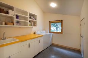 A compact kitchen with a sink, a washing machine, and shelves with various items. - The Gallery House | Architectural Digest–Featured Ketchum Home (Ketchum)
