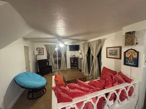 Ein Wohnzimmer mit einem weißen Sofa, einem blauen Sessel und einem roten Schlafsofa. An der Wand befindet sich ein Fernseher und ein Deckenventilator.