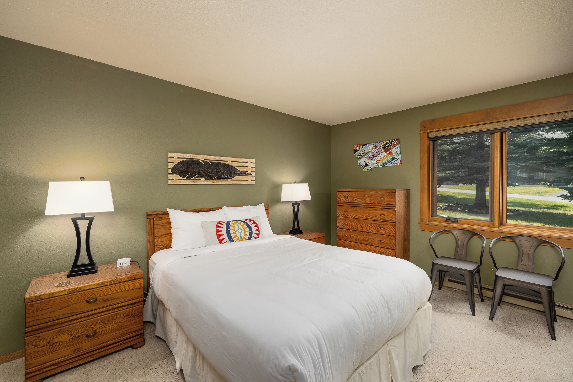 A bedroom with a bed, two nightstands, a dresser, a window with a view, and a wall with a mounted license plate.