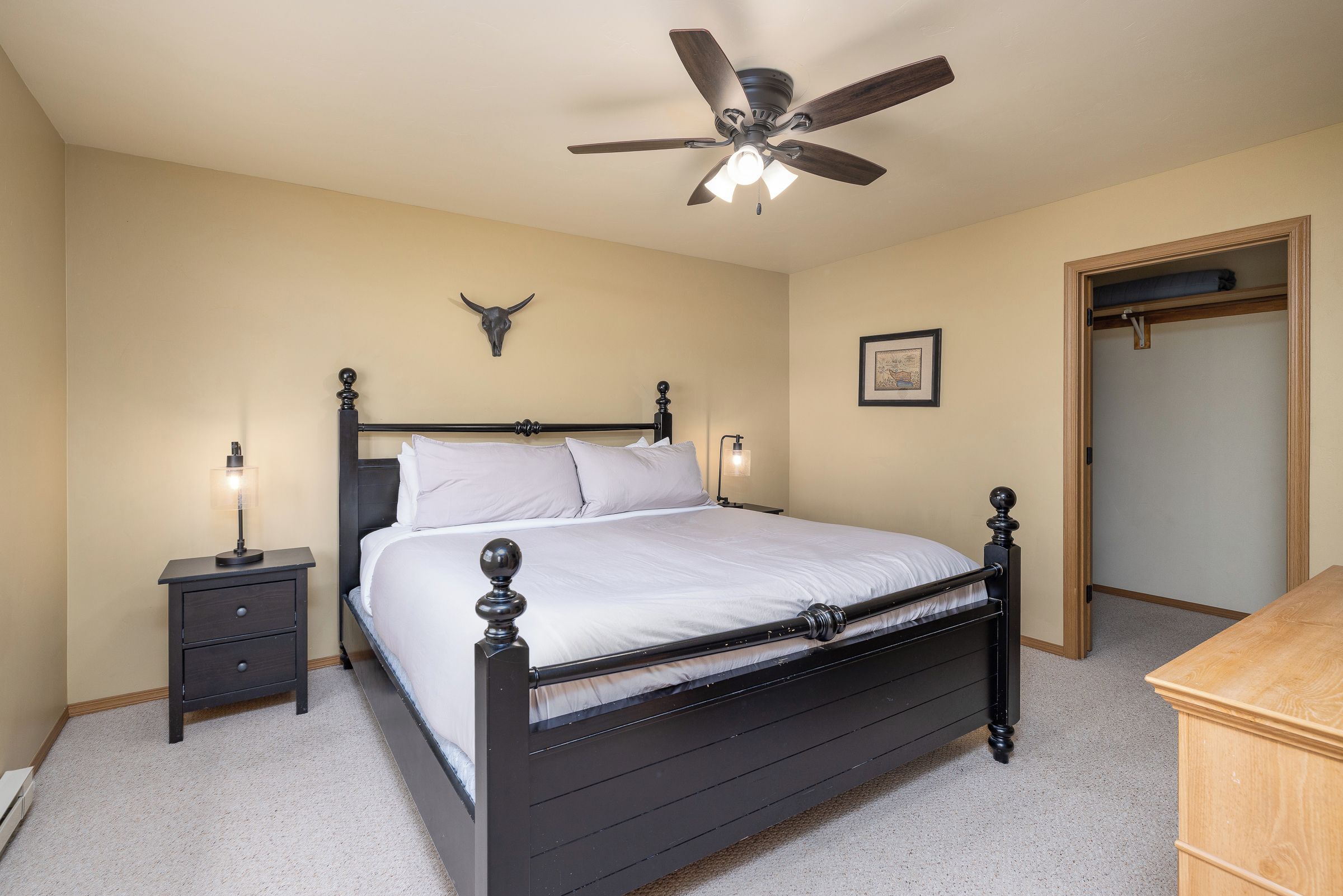 A bedroom with a bed, bedside tables, a ceiling fan, a wall-mounted bull skull decoration, and a framed picture.