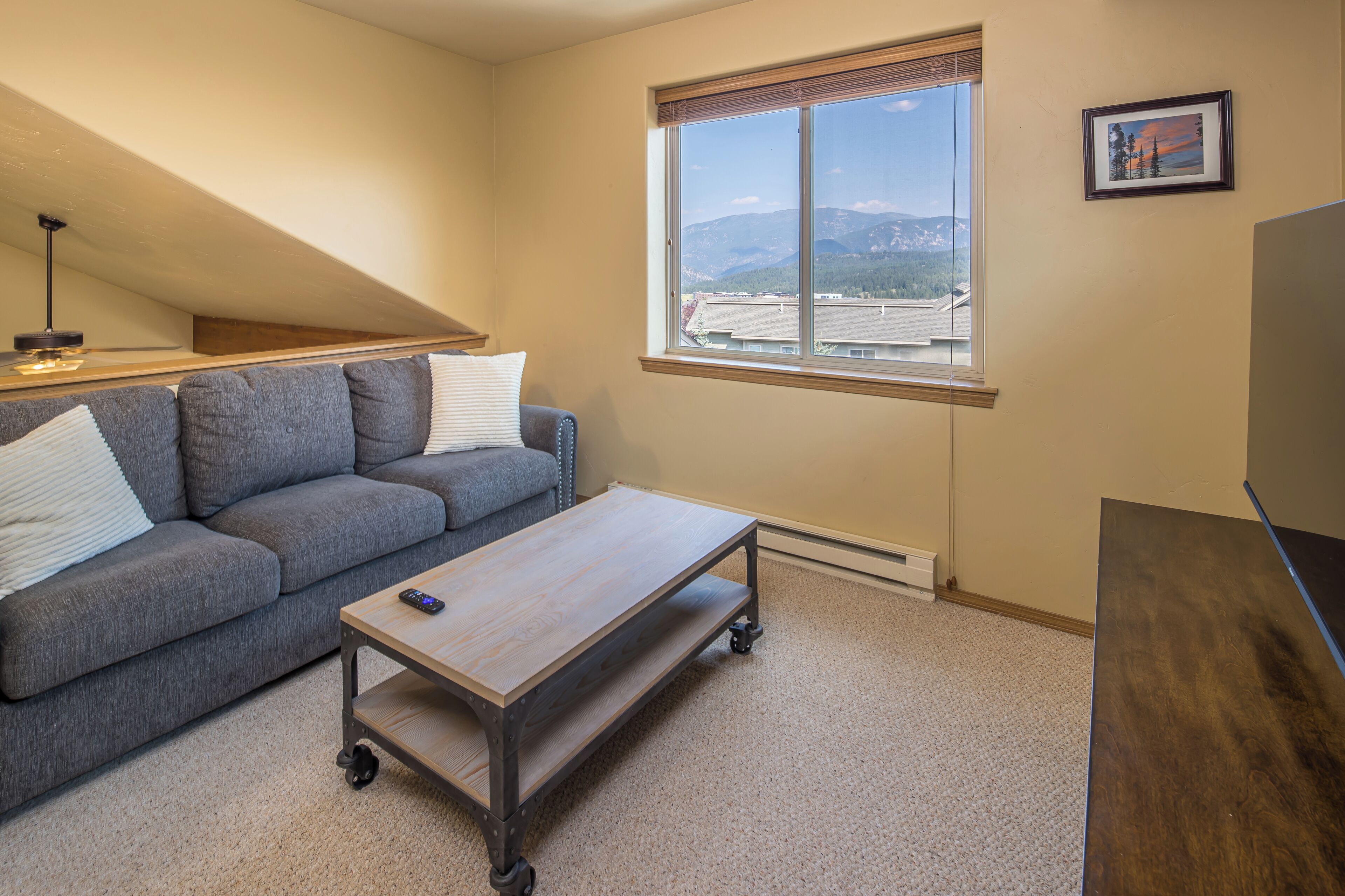 A room with a gray sofa, a wooden coffee table with wheels, a window with a view of buildings and mountains, and a framed picture on the wall.
