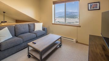 A room with a gray sofa, a wooden coffee table with wheels, a window with a view of buildings and mountains, and a framed picture on the wall.