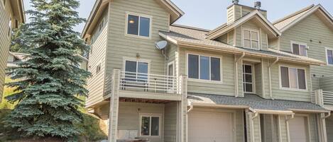 A two-story house with a garage, a balcony, and a large evergreen tree in the front yard.