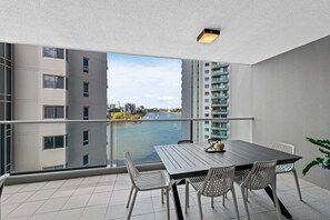 A balcony with a view of a river and buildings, a table with chairs, and a potted plant. - Spacious 3BR | Pool, Gym, Tennis & Parking (Brisbane)