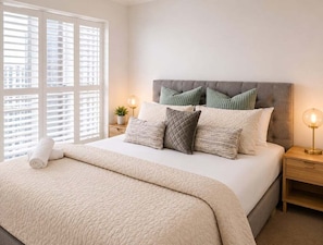 A neatly made bed with a quilted cover, a headboard with multiple pillows, bedside tables with lamps, and a window with blinds. - Spacious 3BR | Pool, Gym, Tennis & Parking (Brisbane)